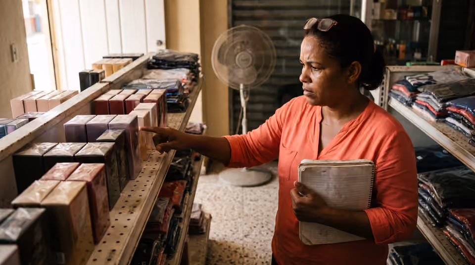 Dueña de una tienda de variedades contando productos en el estante con una libreta apoyada en el antebrazo.