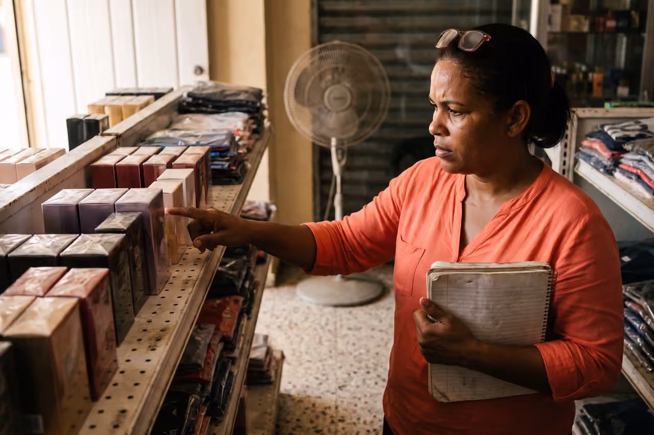 Dueña de una tienda de variedades contando productos en el estante con una libreta apoyada en el antebrazo.