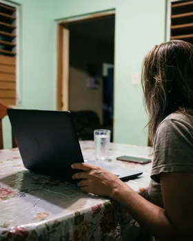 Una mujer sentada a la mesa de la cocina de su casa frente a una computadora portátil, de noche.