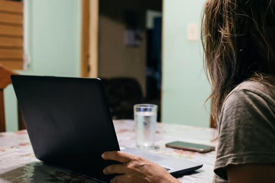 Una mujer sentada a la mesa de la cocina de su casa frente a una computadora portátil, de noche.