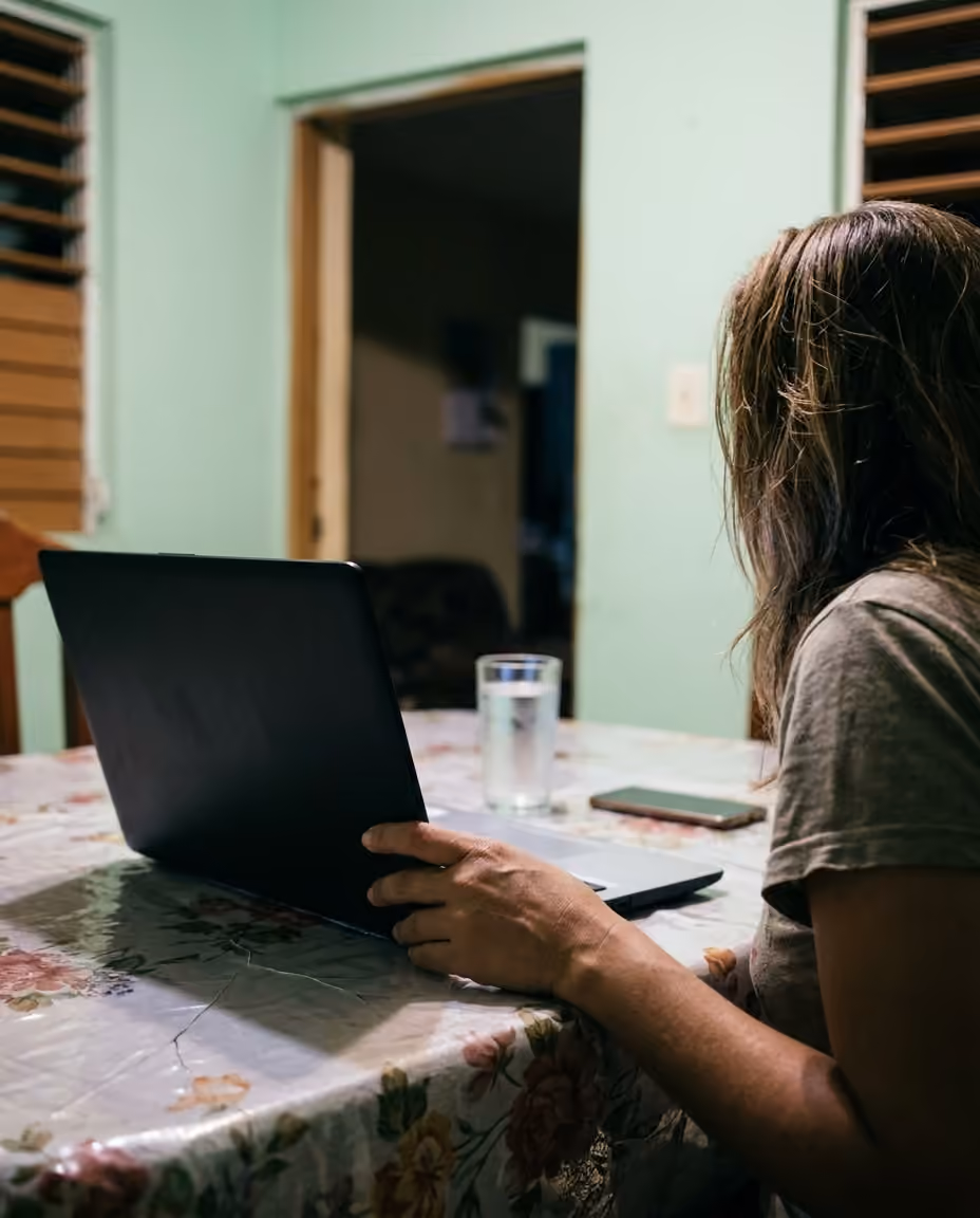 Una mujer sentada a la mesa de la cocina de su casa frente a una computadora portátil, de noche.