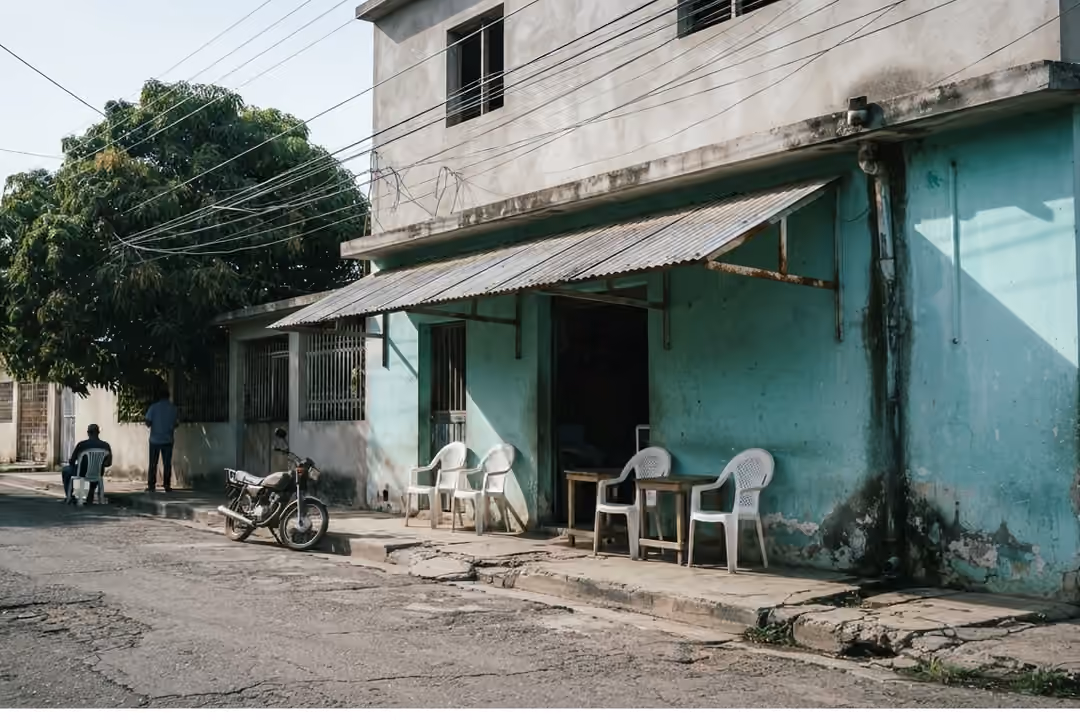 Fachada de un colmado dominicano de esquina al mediodía, con sillas plásticas y un motor parqueado en la acera.